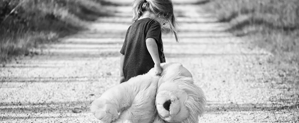 Girl walking on a road carrying a teddy bear.